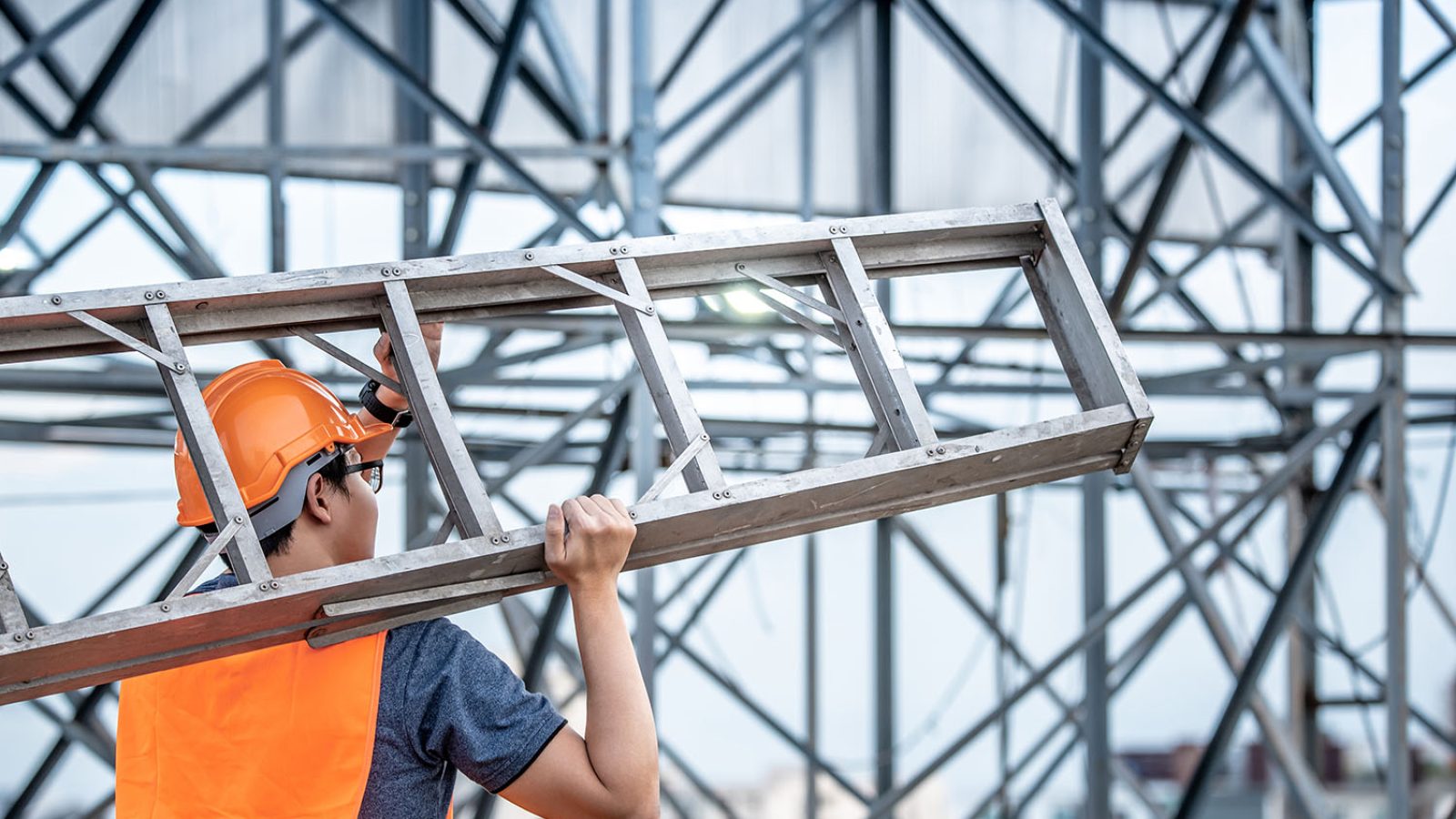 Young Asian maintenance worker with orange safety helmet and vest carrying aluminium step ladder at construction site. Civil engineering, Architecture builder and building service concepts